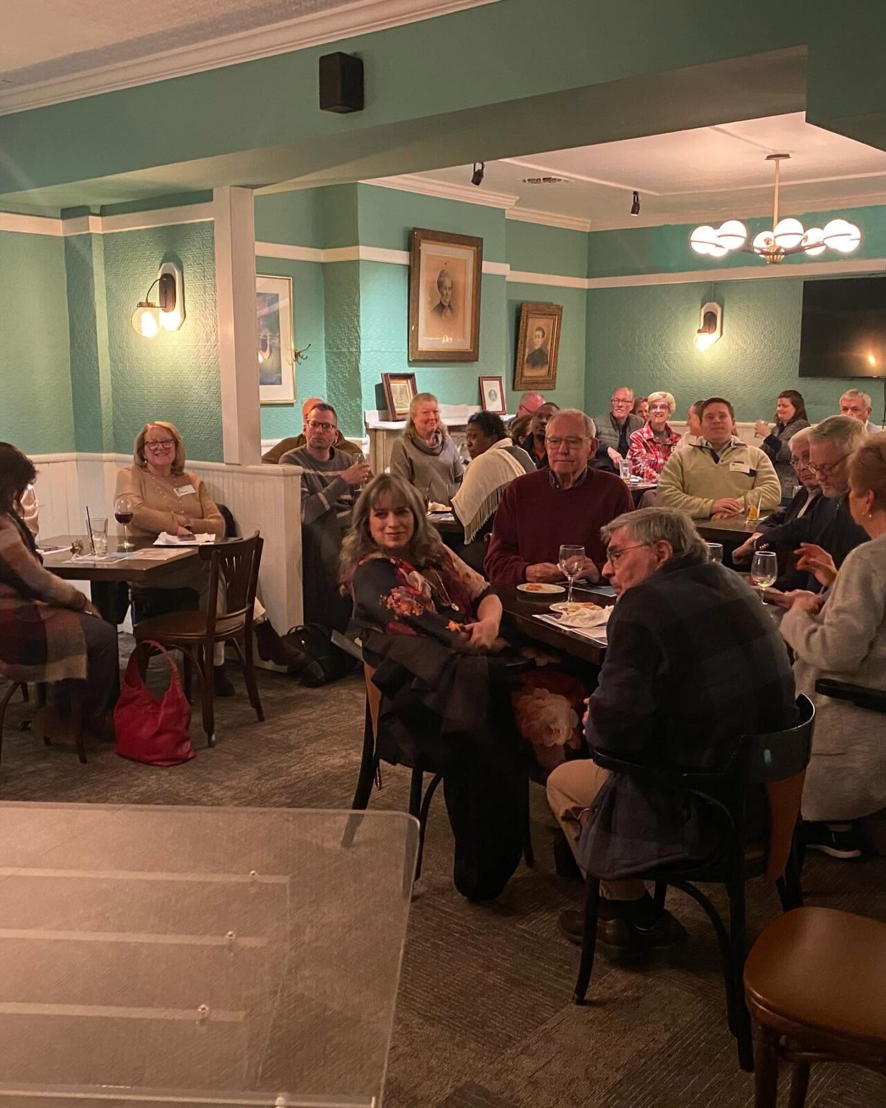 Community Dinner Gathering Woodbury Restaurant Guests sit at tables with wine glasses in a teal-walled restaurant dining room during an evening event.