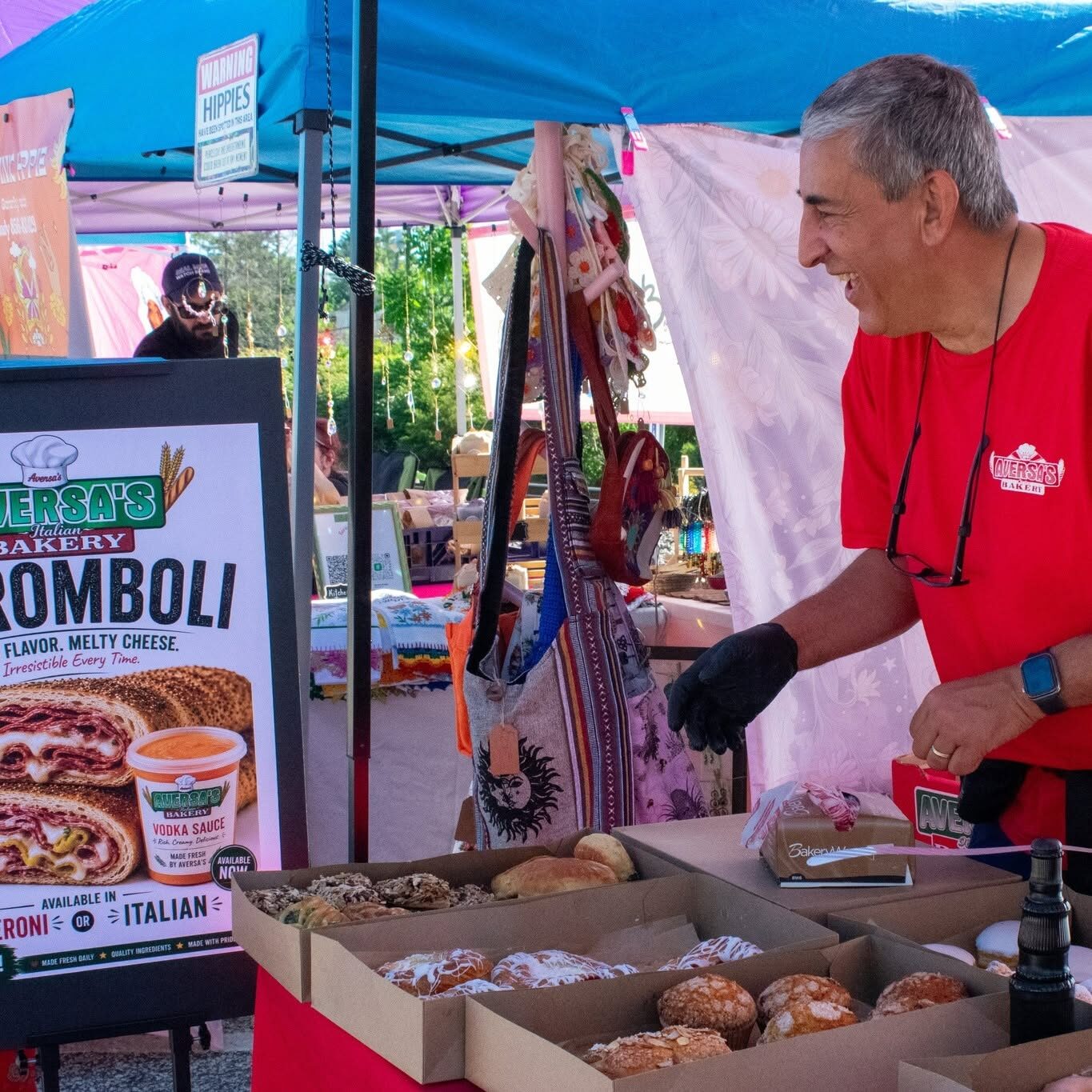 Italian Bakery Vendor Farmers Market A vendor in a red bakery shirt smiles behind open boxes of pastries at a market stall.