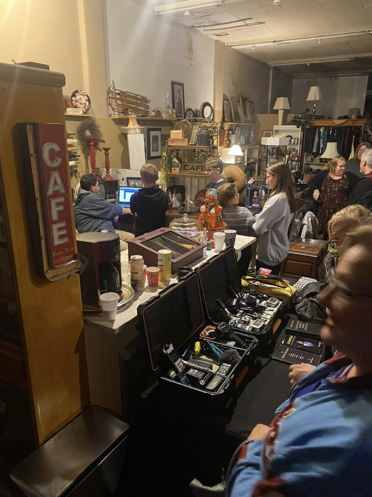 Paranormal Investigation Woodbury Antique Shop People gather among antiques and open cases of ghost-hunting equipment inside a dimly lit shop.