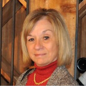 Sandy Uzdavinis Headshot Sandy Uzdavinis smiles in a grey sweater and red turtleneck with a gold necklace.