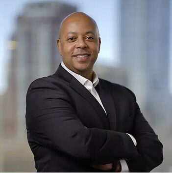 Scott Mccoy Headshot Scott McCoy stands with arms crossed in a dark suit before a blurred city skyline.