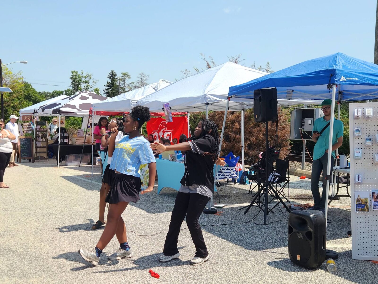 Teen Singers Performing Farmers Market Two teenagers sing and dance with a microphone in front of market tents and a speaker.