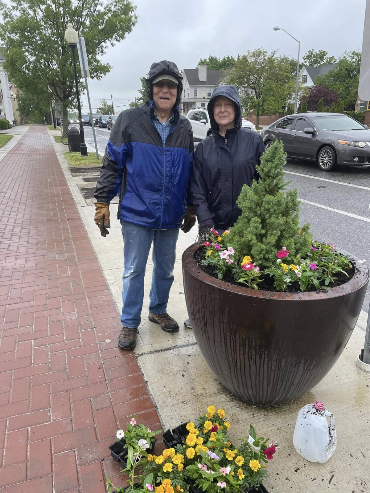 Volunteers Planting Downtown Flower Planter An older couple in rain jackets stand beside a freshly planted sidewalk flower pot on a wet brick walkway.